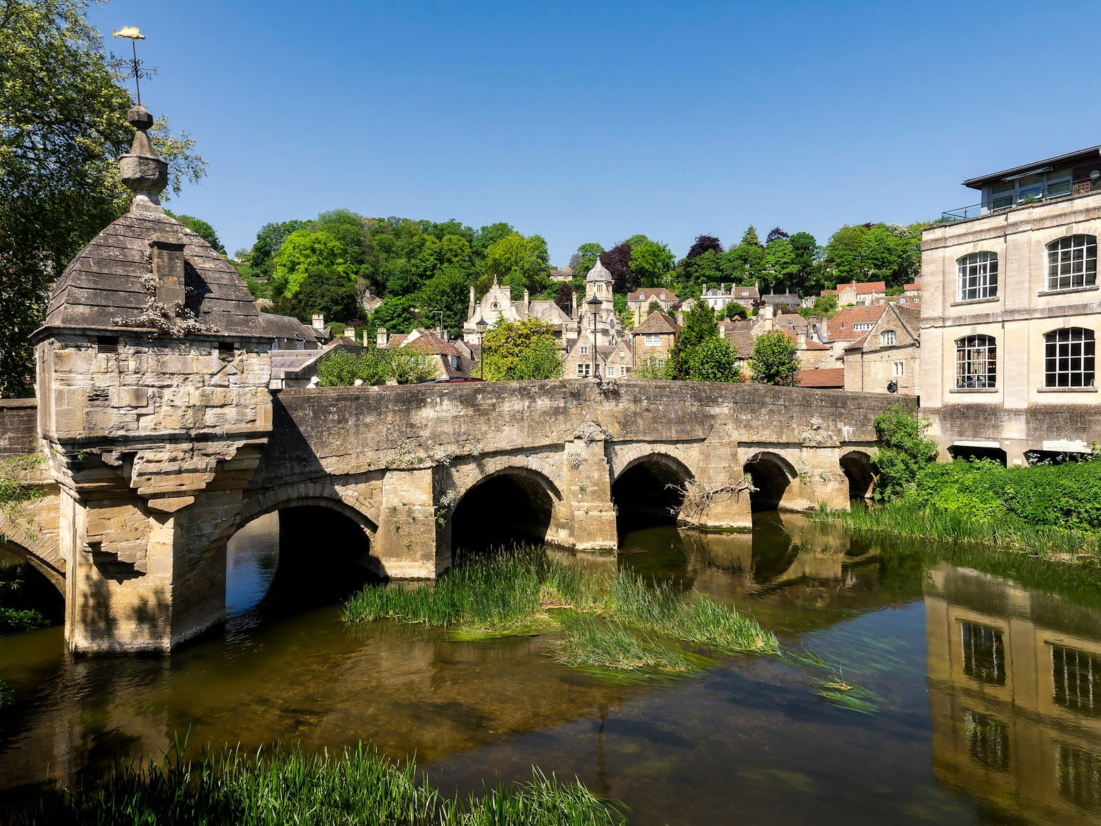 Historic stone bridge over a river in Bradford on Avon, surrounded by lush greenery and traditional buildings, showcasing the picturesque landscape of the area served by Chippenham Cleaning.