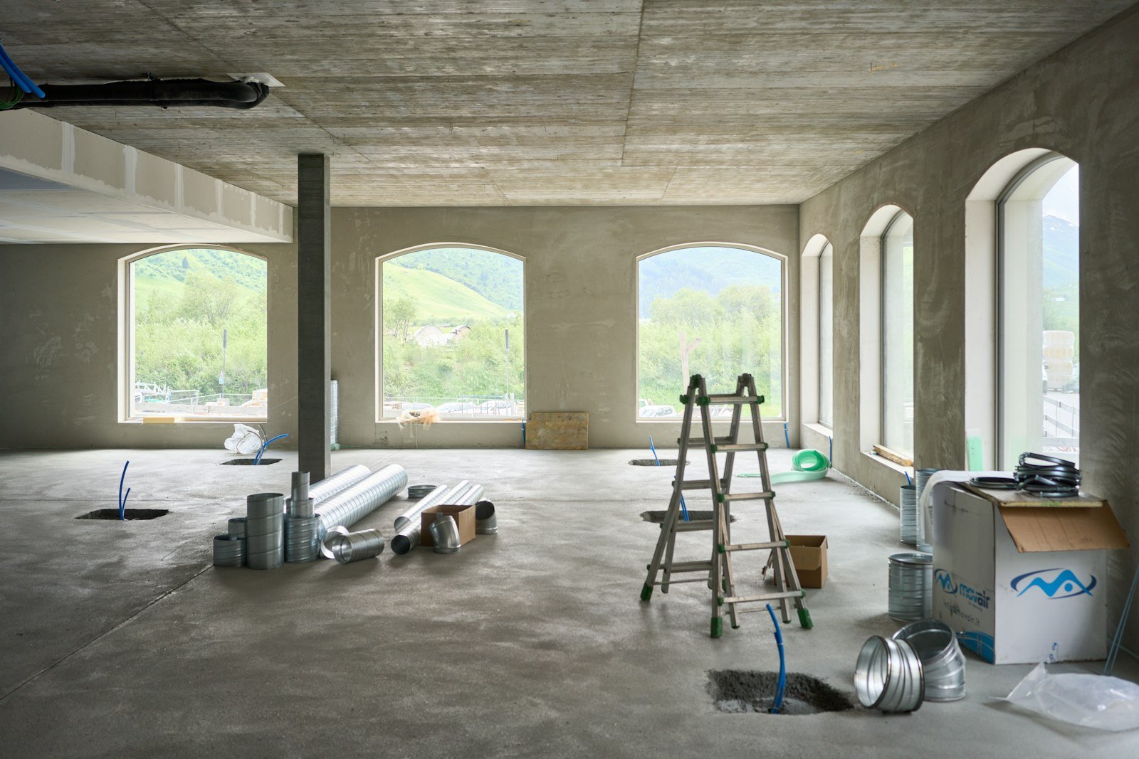 Interior of a new build construction site in Chippenham, featuring a ladder, construction materials like pipes and ducts, unfinished concrete floors, and large windows with views of greenery.